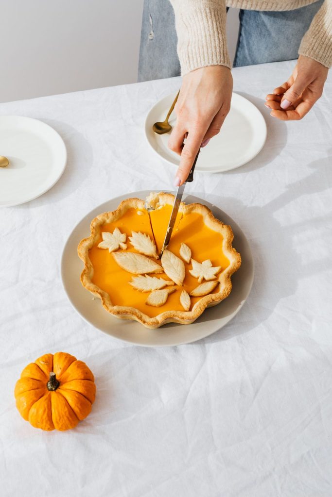 Pumpkin pie with pastry leaf decorations being sliced on a white table beside a small pumpkin, baked ahead