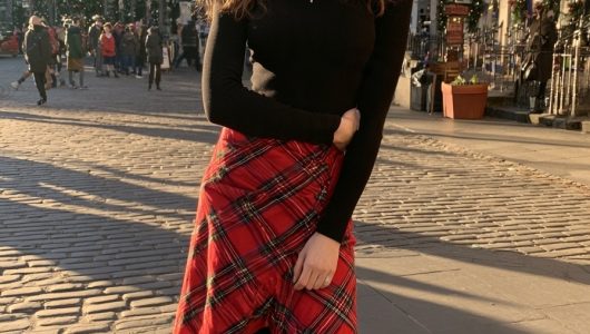 Woman in a red tartan wrap skirt with a black fitted top, black tights and ankle boots on a festive cobbled street