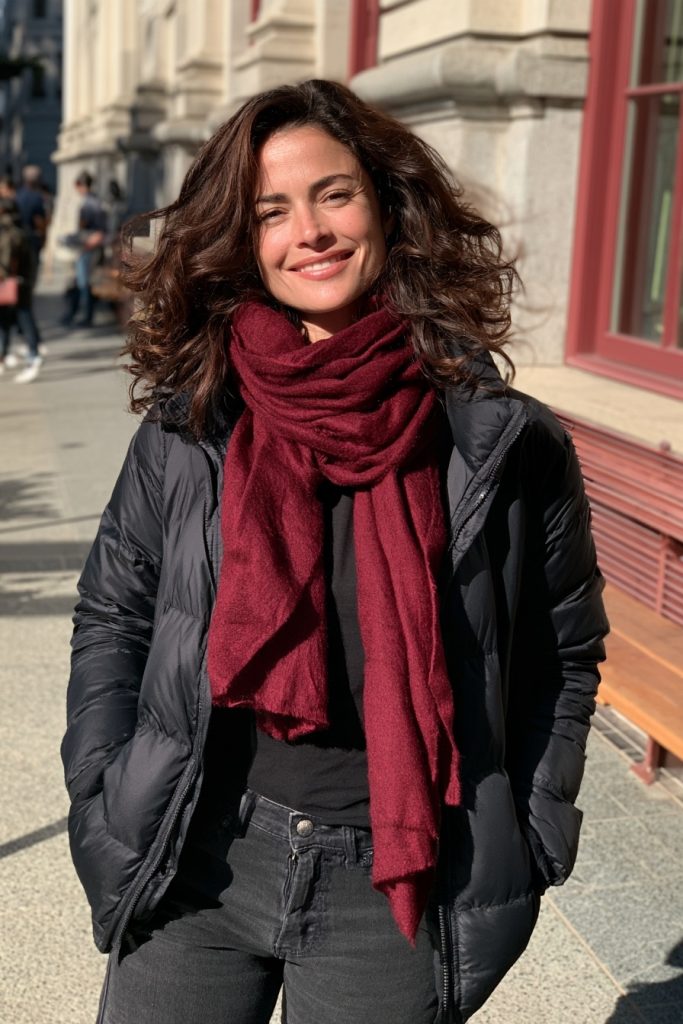 Woman smiling in a burgundy scarf and a black puffer jacket with dark jeans on a city street with curly hair