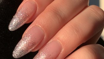 Close-up of a hand with silver glitter ombre almond nails fading from a sheer pink base on black velvet