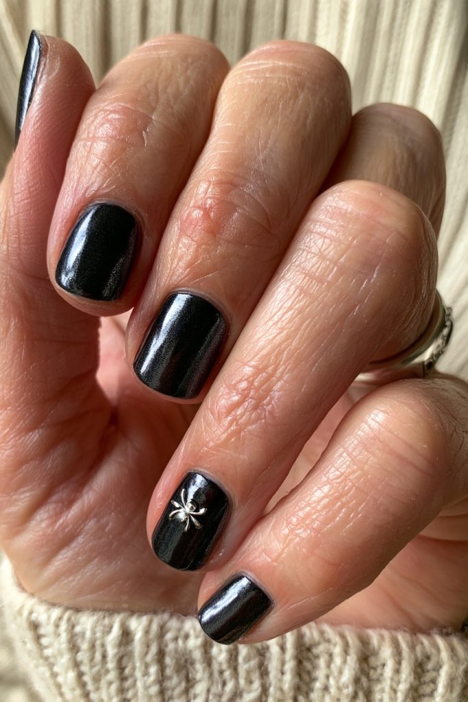 Hand with short black chrome nails and a tiny silver spider charm on one nail