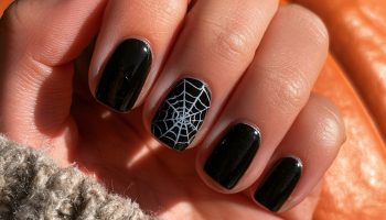 Hand with short black nails and a white spider web design on the ring finger resting on an orange pumpkin