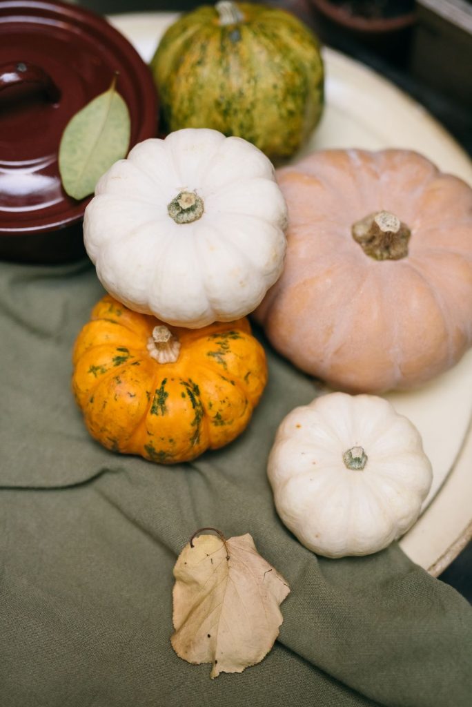 White, orange and blush pumpkins stacked on a sage green cloth runner with dried leaves for a fall table