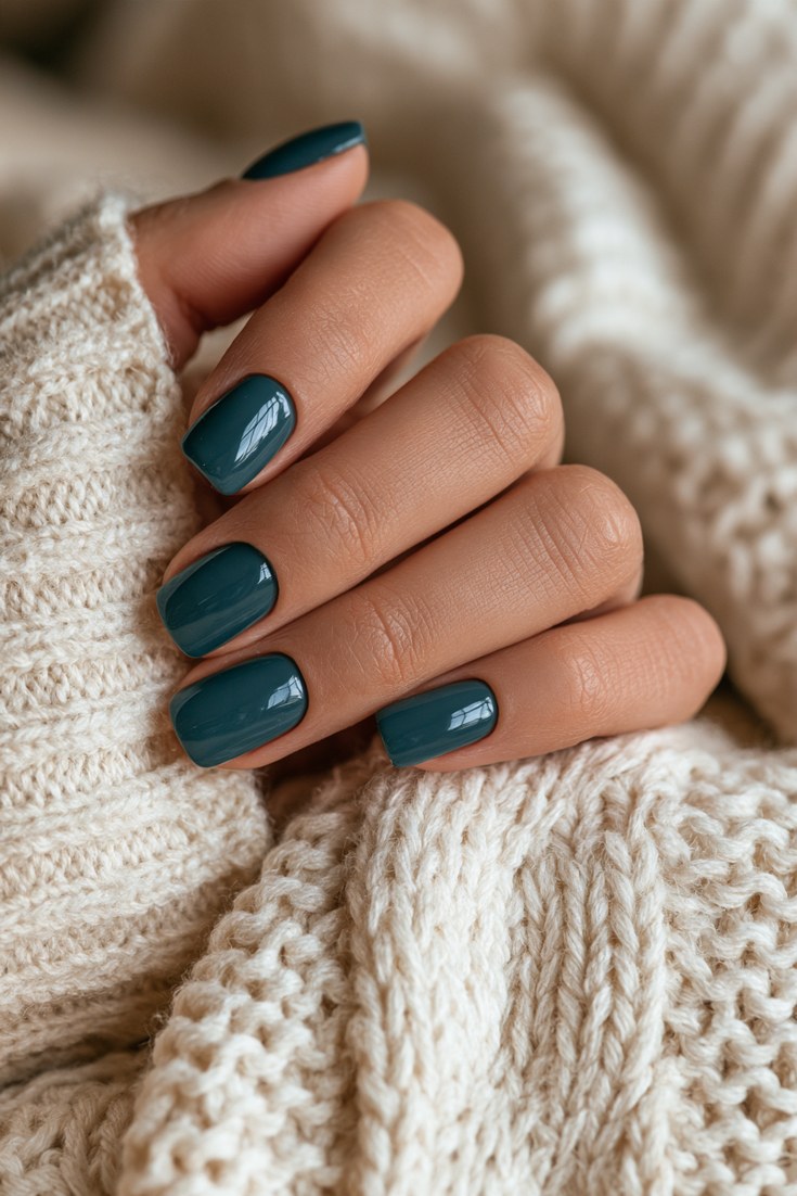 Short square nails in deep teal cream gel on a hand resting on a cream knit blanket