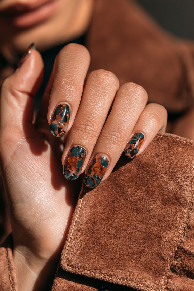 Medium oval nails with a teal and amber tortoiseshell swirl on a hand resting on a brown suede jacket