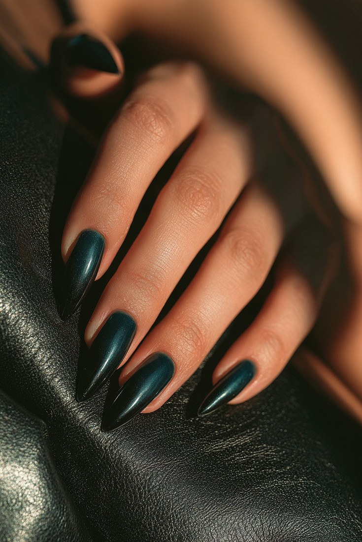 Long stiletto nails in a glossy deep teal to black ombre on a hand resting on black leather