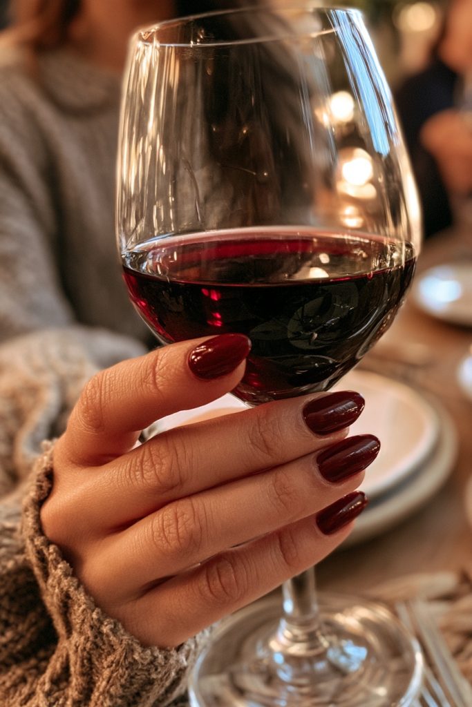 Hand with long almond nails in deep burgundy wine polish holding a stemmed glass of red wine at a candlelit table