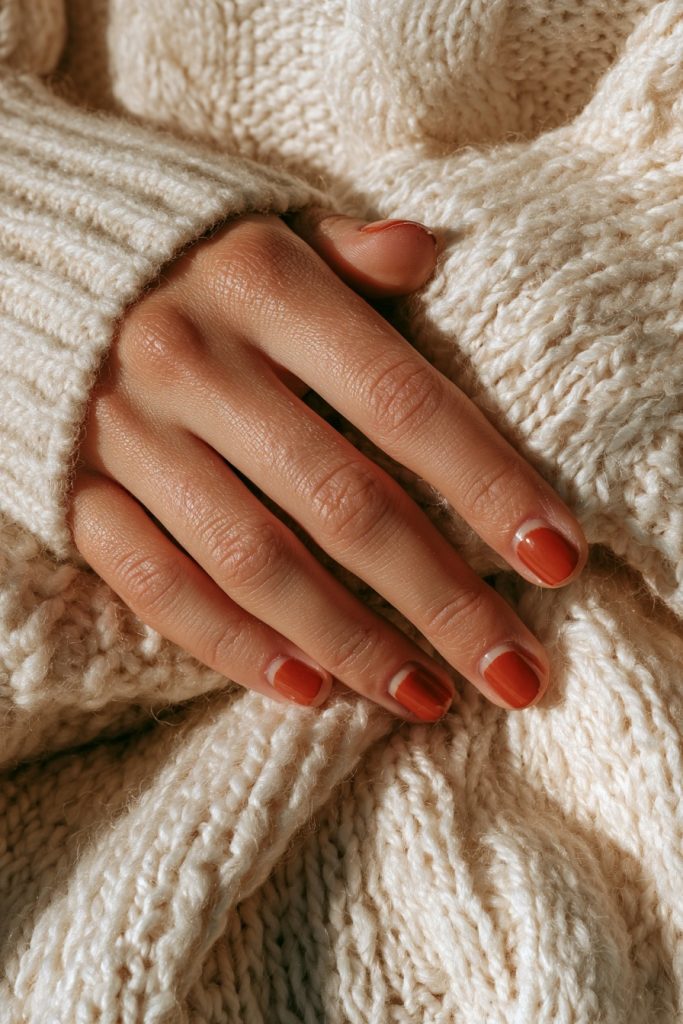 Hand with short natural nails and thin rust red French tips resting on a cream knit sweater