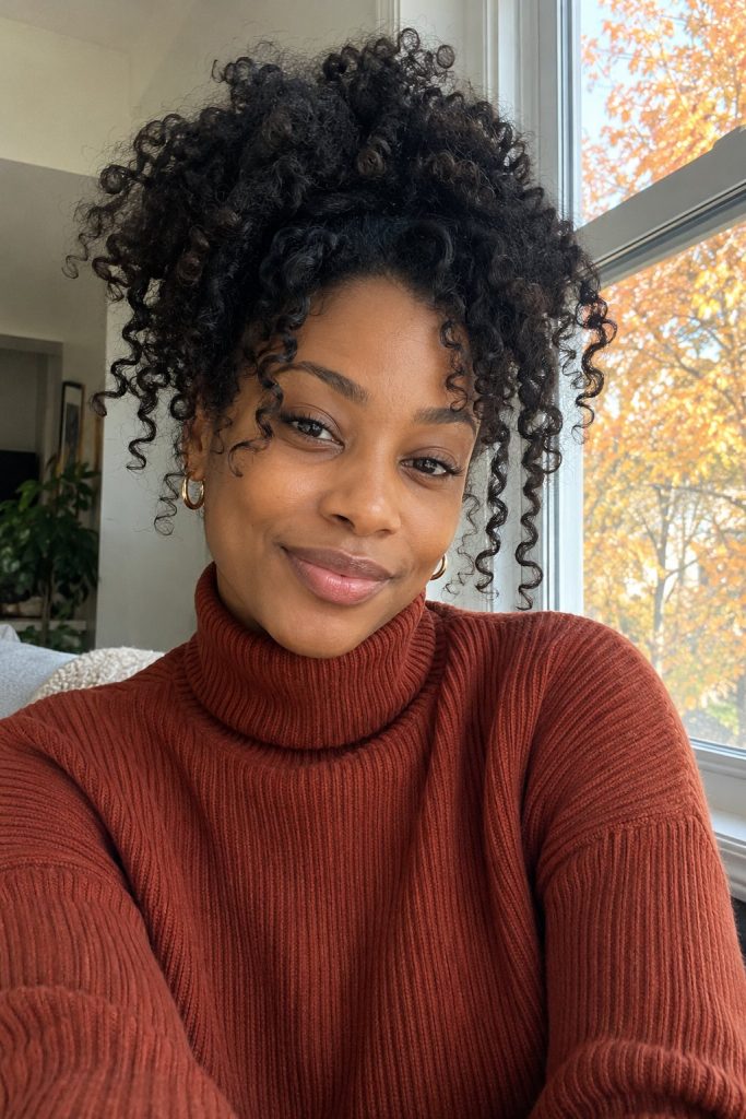 Coily curls gathered in a high pineapple puff with a rust ribbed turtleneck
