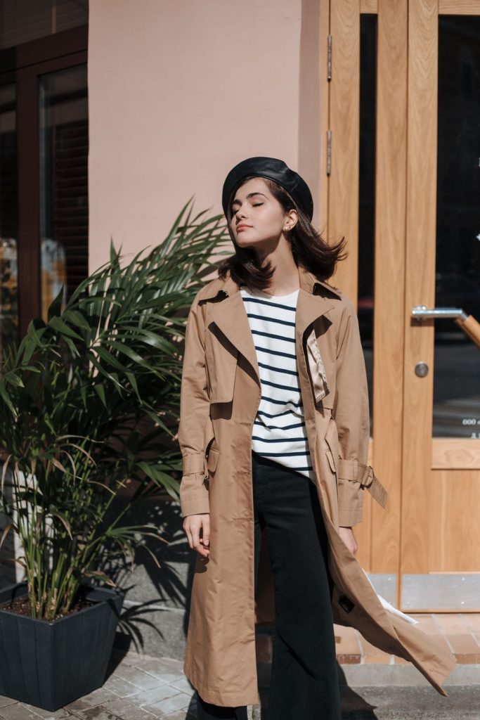 Woman in a camel trench coat over a black and white striped top with black trousers and a black beret