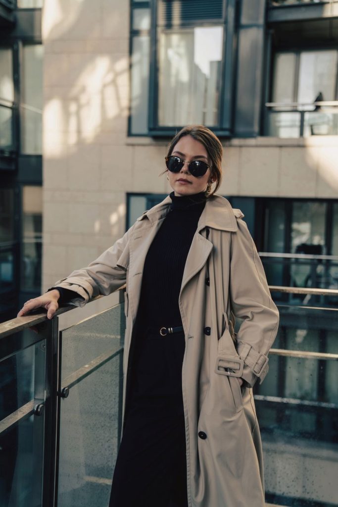 Woman in sunglasses wearing a beige trench coat over a black turtleneck beside a glass railing
