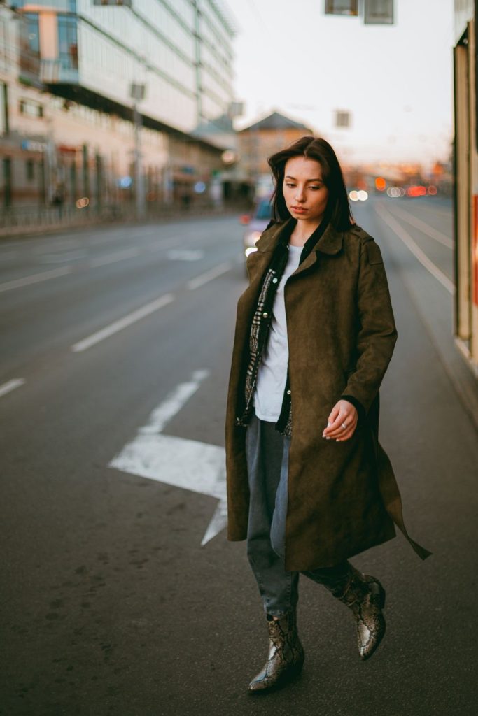 Woman crossing a city street in an olive green trench-style coat, a white shirt, dark jeans and black boots