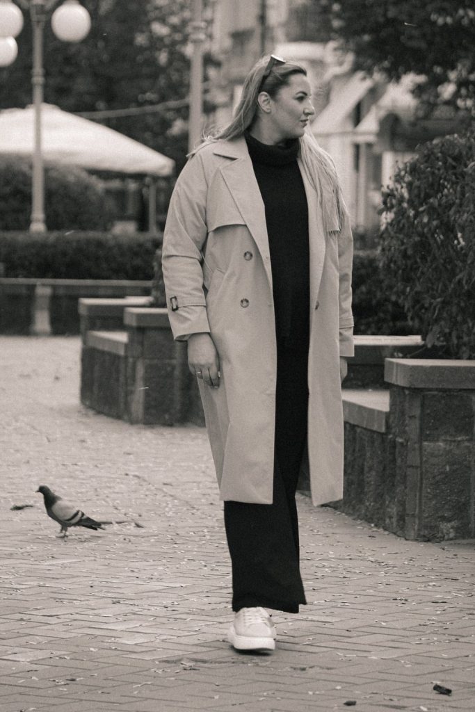 Woman in a pale grey-beige trench coat over a black turtleneck and long black skirt with white sneakers