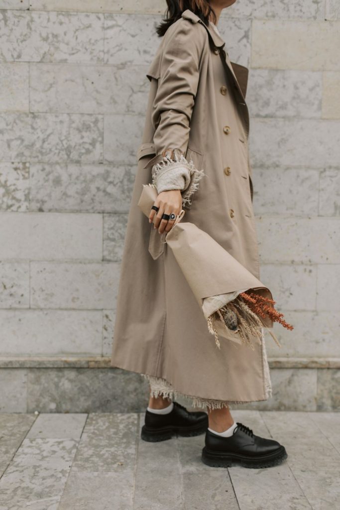 Woman in a long beige trench coat holding a bouquet of dried flowers, with white socks and chunky black lace-up shoes