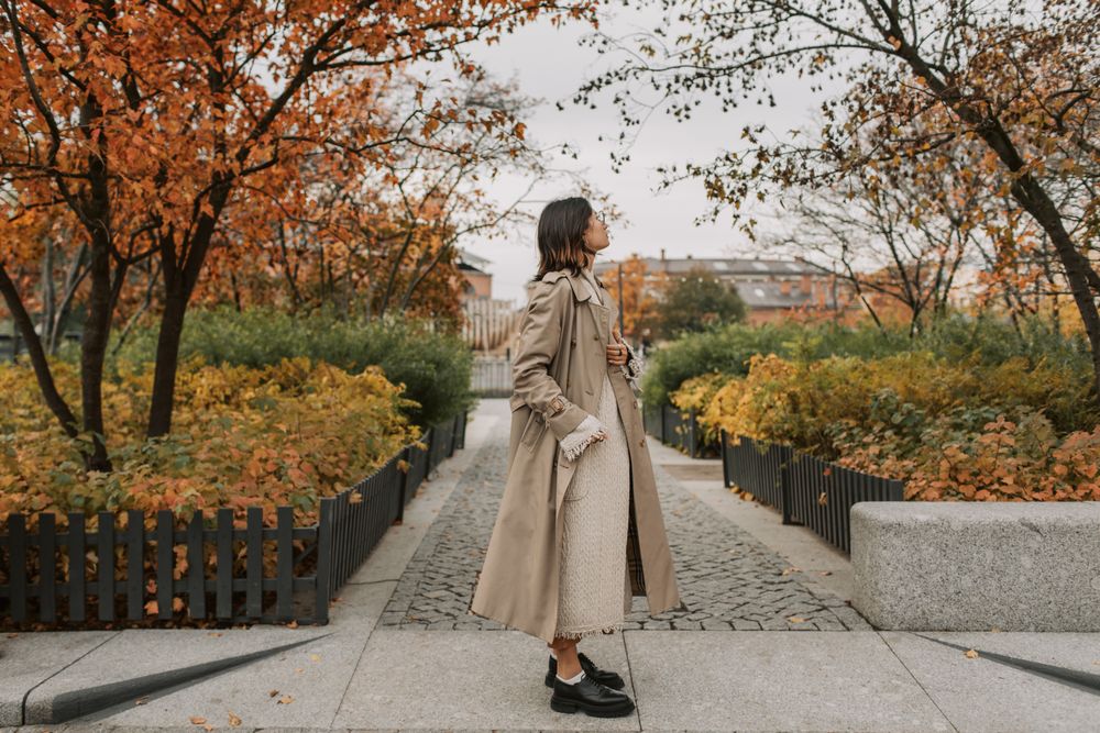 Woman on a cobblestone path in a long beige trench coat with chunky black shoes among autumn trees