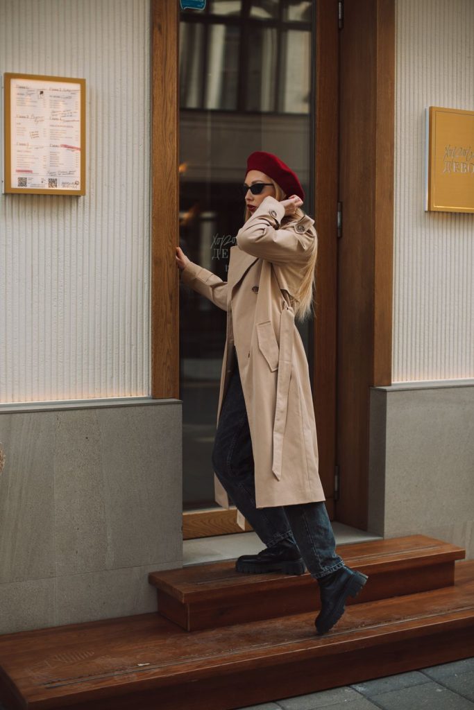 Woman in a beige trench coat, a red beret and sunglasses stepping out of a cafe doorway with dark trousers