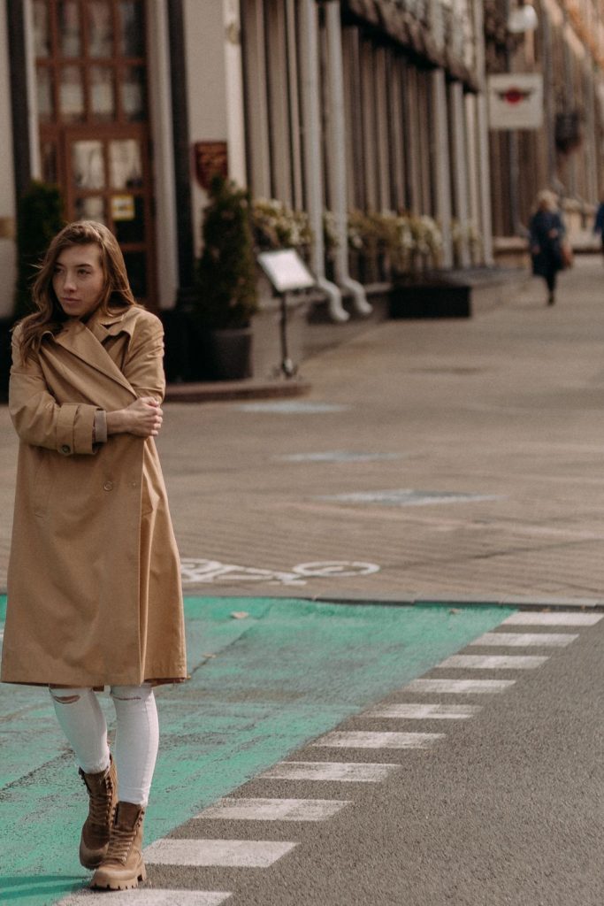 Woman in a long belted camel coat with white jeans and brown ankle boots crossing a street