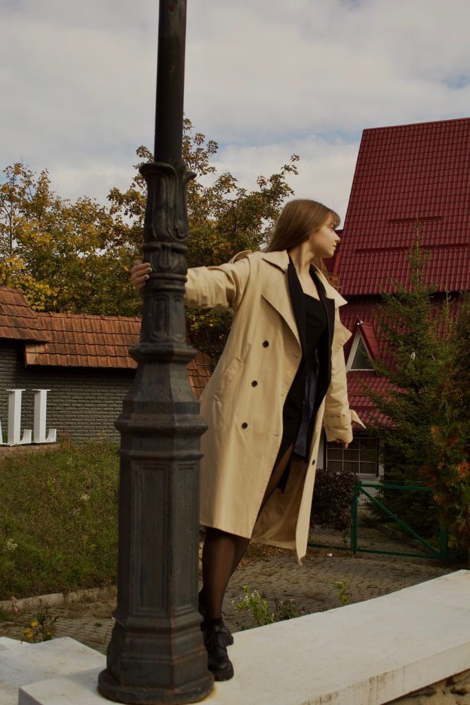 Woman leaning on a lamppost in an open tan trench coat over a black dress and black tights