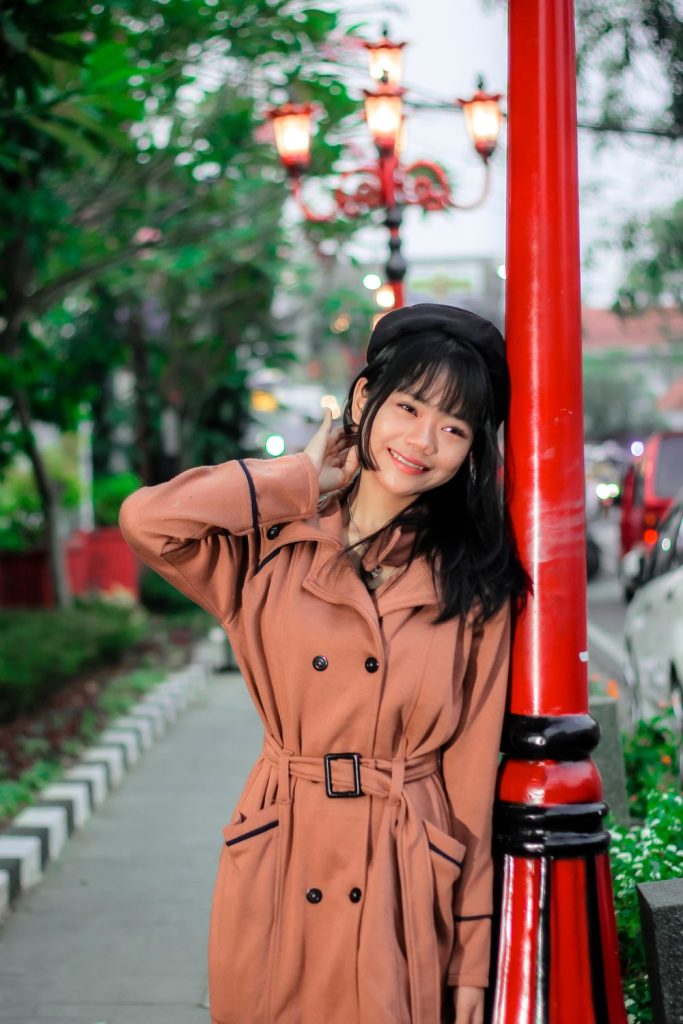 Smiling woman in a rust orange double-breasted belted trench coat and a black beret leaning on a red lamppost