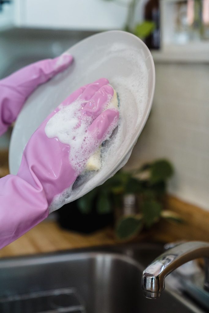 Hands in pink rubber gloves washing a white plate with soapy foam at the kitchen sink