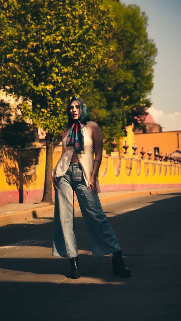 Woman with a blue headscarf in a cream waistcoat and grey-blue wide leg jeans standing in front of a yellow wall