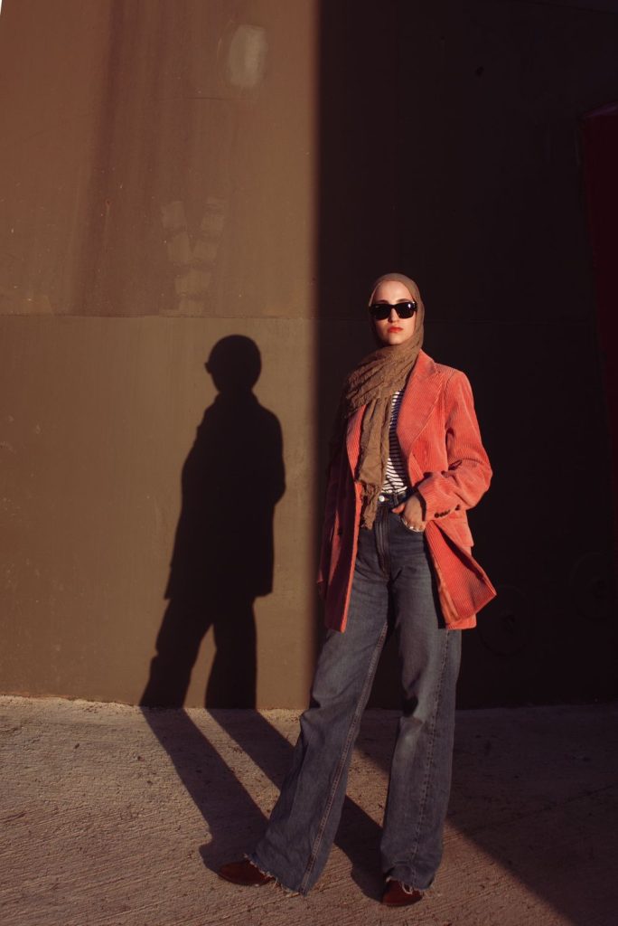 Woman in sunglasses and a rust orange blazer with a scarf and dark grey wide leg jeans standing against a wall with a long shadow