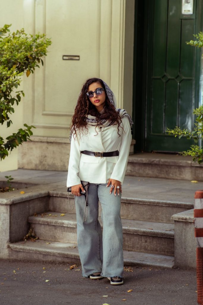 Woman with curly hair and sunglasses in a white jacket with a black belt, a patterned scarf and light grey wide leg jeans on stone steps