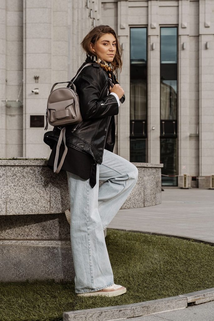 Woman in a black leather jacket with a grey backpack and very light wide leg jeans leaning on a stone ledge