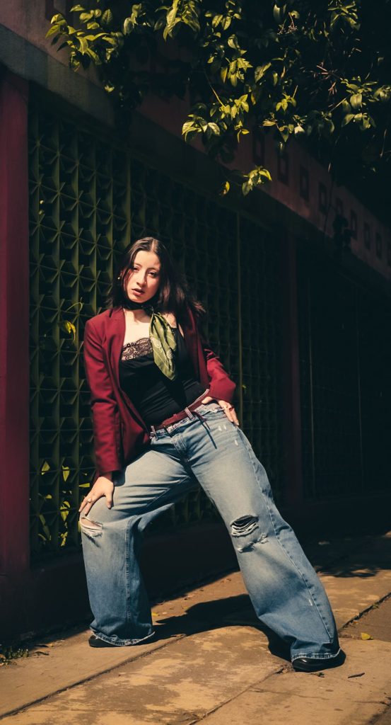 Woman in a burgundy blazer, a black lace-trimmed top, a belt and distressed wide leg jeans with a green scarf at the neck