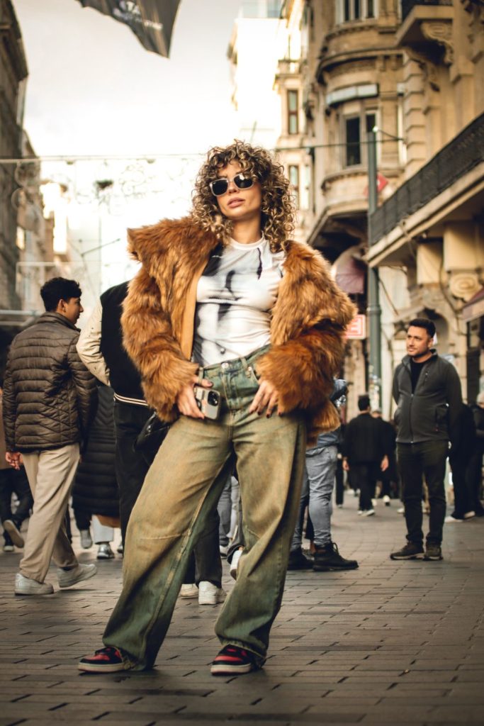 Woman with curly hair and sunglasses in a brown faux fur jacket, a graphic tee and khaki wide leg trousers in a busy street