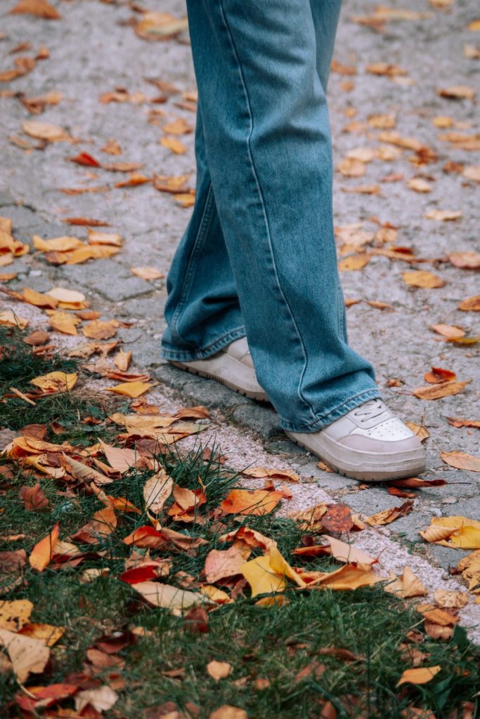Close-up of a wide leg blue jean and white sneaker on a pavement covered with orange and yellow autumn leaves