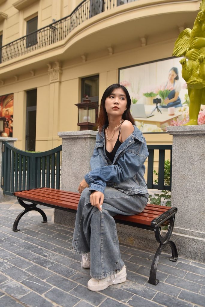 Woman sitting on a red bench in a washed denim jacket worn off the shoulder over a black top with grey-blue wide leg jeans