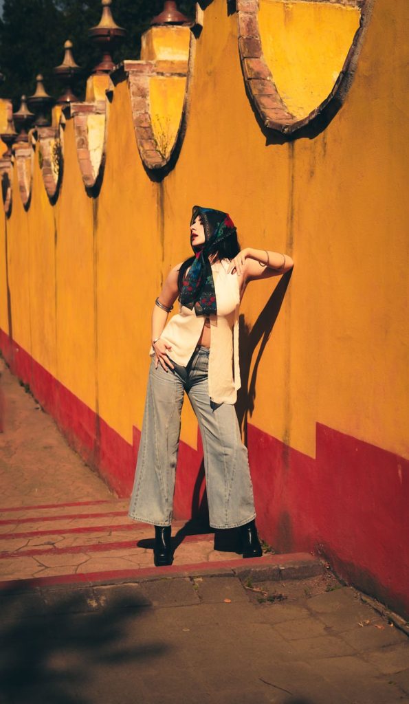 Woman in a headscarf, a cream waistcoat and grey wide leg trousers with black boots in front of a yellow wall