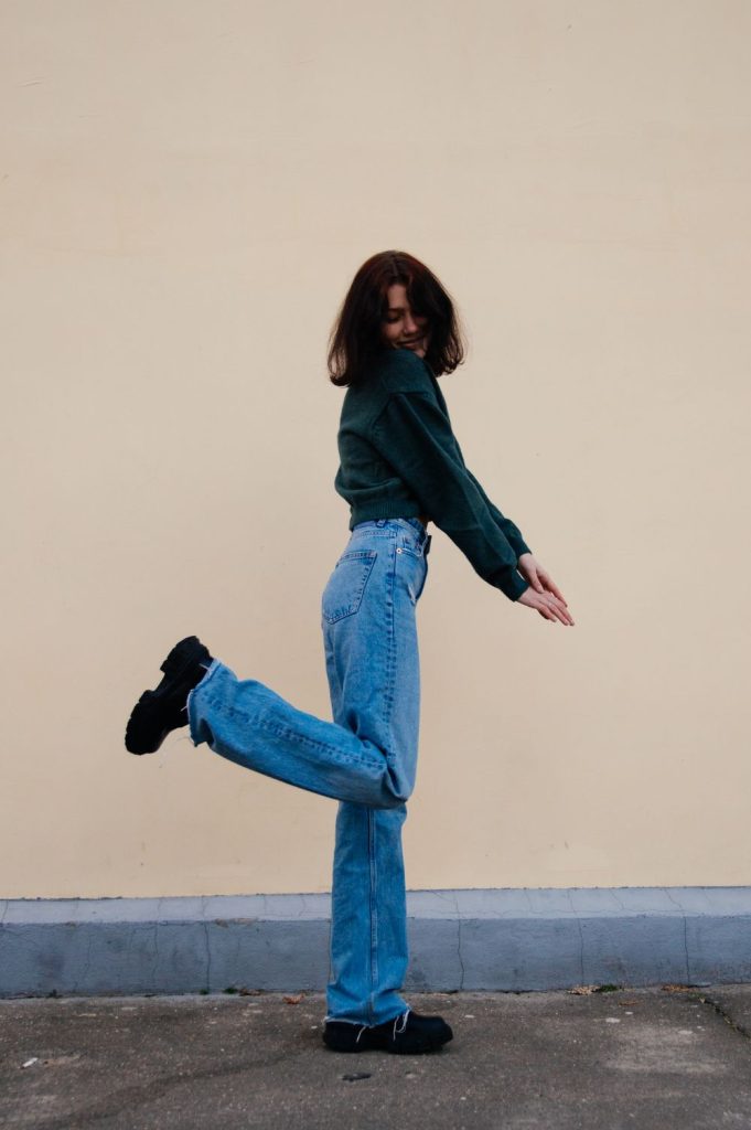 Woman in a dark green sweater and mid-wash wide leg jeans kicking one leg up with a chunky black shoe