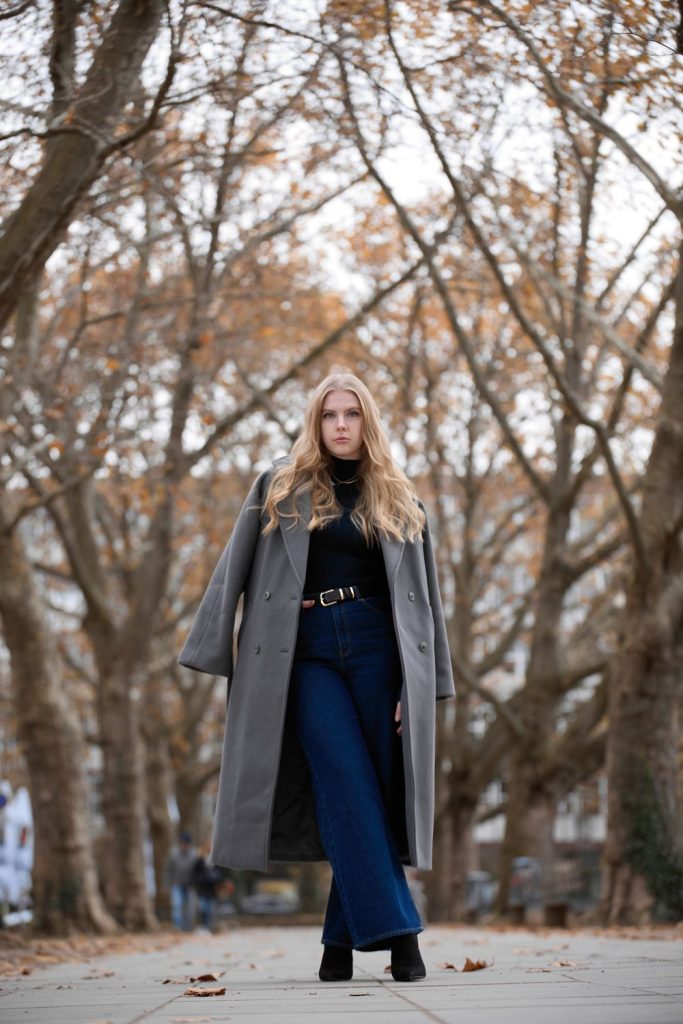 Woman with long blond hair in a long grey coat, a black turtleneck, a belt and dark navy wide leg jeans standing among bare autumn trees