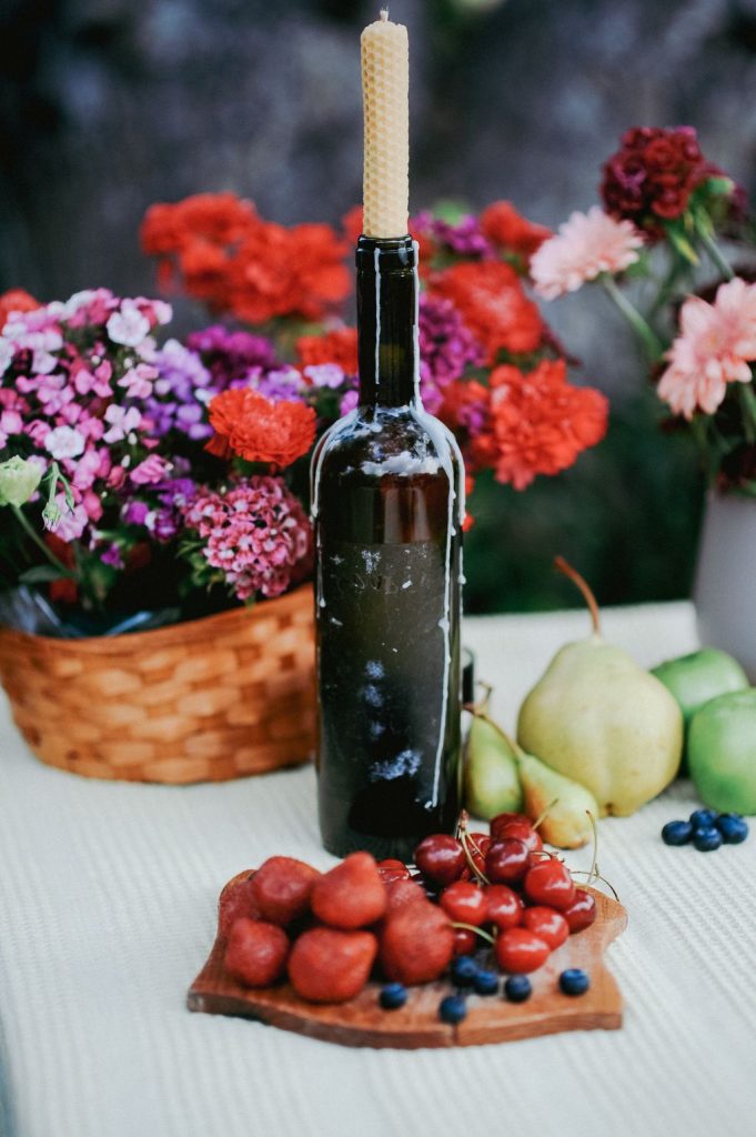Dark wine bottle used as a candle holder with a cream taper, beside pears, berries and a basket of flowers