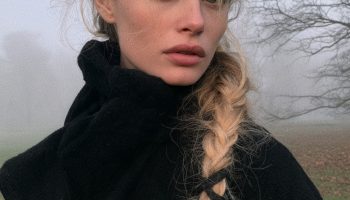 Woman with a long side braid threaded with black ribbon and small silver charms wearing a black coat in a misty park